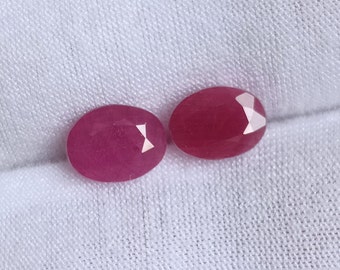 Puede incluir: Dos piedras preciosas de rubí de forma ovalada y facetadas, ambas de color rosa intenso, se muestran sobre un fondo de tela blanca.