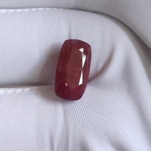 Pode incluir: Uma pedra preciosa vermelha escura, facetada e de formato retangular. A gema tem uma superfície polida com múltiplas facetas que refletem a luz. A pedra está sobre um fundo de tecido branco, destacando sua cor e clareza. Esta gema pode ser usada em joias.