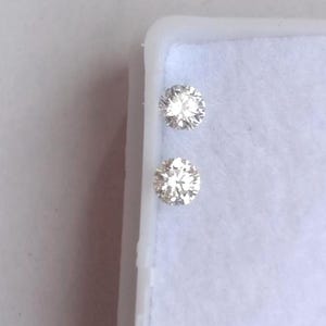May include: Two round-cut diamonds, each with multiple facets, are displayed against a white background. The diamonds sparkle, reflecting light, and are set in a white jewelry box. The gems are a light, clear color.