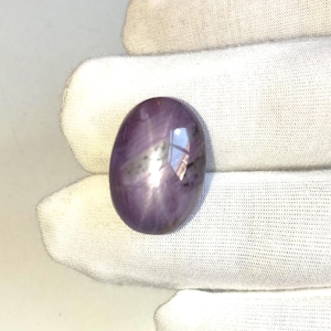 Könnte beinhalten: Ein polierter, oval geformter, violetter Edelstein. Der Edelstein hat eine glatte Oberfläche mit subtilen inneren Mustern und einem reflektierenden Glanz. Der Stein ist vor einem weißen Hintergrund platziert, der seine Farbe und Form hervorhebt. Der Edelstein ist im Cabochon-Schliff.