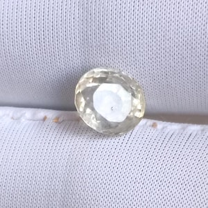 May include: A faceted, oval-cut gemstone with a pale yellow hue. The gem is set against a white fabric background, showcasing its clarity and brilliance. The gemstone's facets reflect light, creating a sparkling effect.