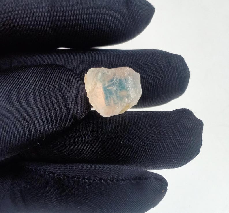 Puede incluir: Una peque&ntilde;a piedra preciosa blanca transl&uacute;cida con una inclusi&oacute;n azul y verde. La piedra preciosa se est&aacute; sosteniendo con un guante negro.