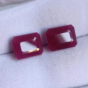 Pode incluir: Duas pedras preciosas retangulares facetadas de cor vermelha com um tom vermelho ligeiramente mais escuro no centro. As pedras preciosas estão sobre uma superfície de tecido branco.