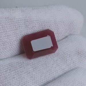May include: A deep red gemstone with a rectangular shape and a polished finish. The gemstone is set on a white fabric background.
