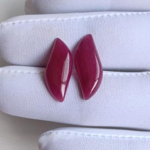 May include: Two polished, teardrop-shaped gemstones in a deep magenta hue. The gems have a smooth, glossy surface, reflecting light. The background is a white, textured fabric.