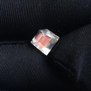 May include: A square-cut gemstone with a clear, faceted exterior and a soft pink interior. The gem is set against a dark, textured background, highlighting its brilliance and color.