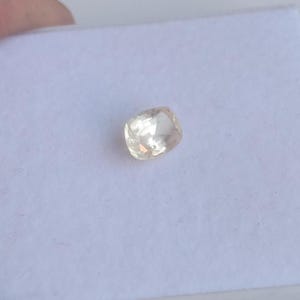 May include: A cushion-cut, light peach-colored gemstone. The gem is set against a white background, showcasing its facets and clarity. The stone has a soft, warm hue, reflecting light.