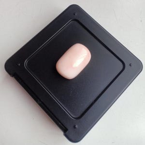 May include: A pale pink oval-shaped gemstone displayed on a black square background.