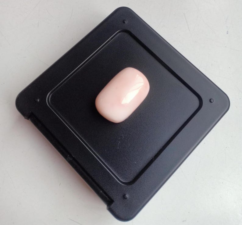 May include: A pale pink oval-shaped gemstone displayed on a black square background.