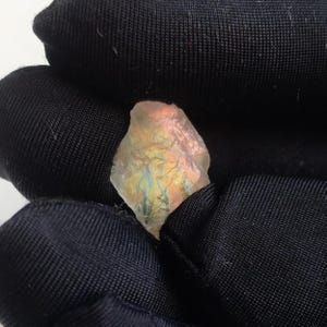 May include: A small, irregularly shaped, translucent gemstone with iridescent colors of orange, yellow, and blue. The stone is held in a black fabric, creating a contrast that highlights the gem's natural beauty. The gemstone appears to be raw and uncut.