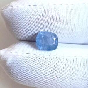 May include: A light blue, cushion-cut gemstone with a faceted surface. The gem is set against a white, textured background. The stone's color is a soft, pale blue, and it appears to be transparent.