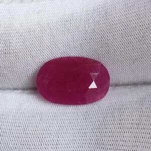 May include: A vibrant pink oval-shaped gemstone with a faceted cut. The gemstone is set against a white fabric background.