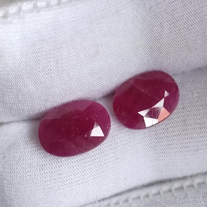 May include: Two oval-shaped, faceted ruby gemstones, set against a white fabric background.