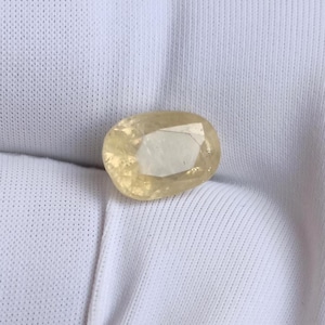 May include: A faceted, oval-shaped gemstone with a pale yellow hue. The gem is set against a white fabric background, highlighting its clarity and color. The cut of the stone reveals multiple facets.