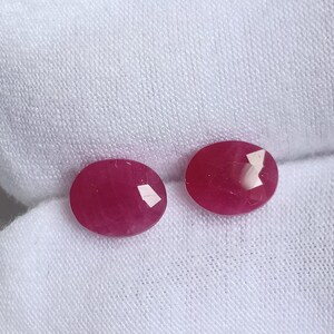 Op de afbeelding: Twee ovale, gefacetteerde robijn edelstenen met een diepe roze tint. De edelstenen liggen op een witte stoffen achtergrond.