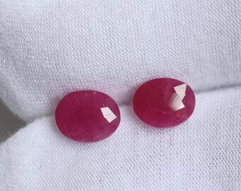 Puede incluir: Dos piedras preciosas de rubí de forma ovalada y facetadas, de un tono rosa intenso. Las piedras preciosas están colocadas sobre un fondo de tela blanca.