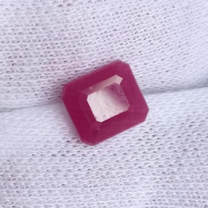 Pode incluir: Uma gema quadrada de cor rosa escura com corte facetado. A gema está sobre um fundo de tecido branco.