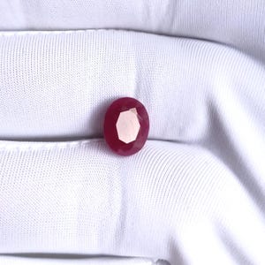 May include: A faceted, oval-shaped ruby gemstone. The gem is a deep red color with a lighter, almost white, interior. The ruby is set against a white, textured fabric background.