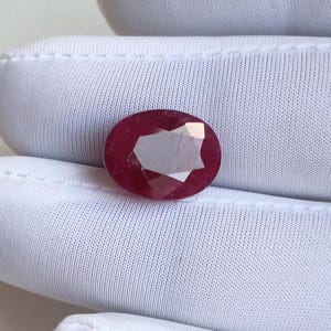 May include: A faceted, oval-shaped ruby gemstone. The ruby is a deep red color with a polished surface, reflecting light. The gemstone is set against a white background, highlighting its color and clarity. This gemstone is suitable for jewelry making.