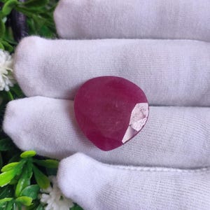 Peut inclure: Une pierre précieuse en forme de cœur, facettée, de couleur rouge rubis. Le rubis est d'un rouge profond et riche avec une surface polie. La pierre est présentée sur un fond de tissu blanc avec du feuillage vert et des fleurs blanches.