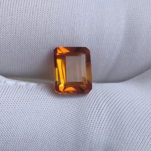 May include: A rectangular, faceted gemstone in a warm amber-orange colour. The gem is set against a white, textured fabric, highlighting its clarity and brilliance. The cut of the stone is precise, showcasing its internal structure and light refraction.