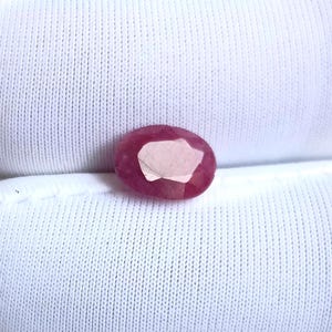 May include: A faceted, oval-shaped ruby gemstone. The ruby is a deep red color with a lighter, irregular-shaped area in the center. The gemstone is set against a white, textured fabric background.