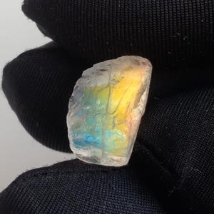 May include: A raw, translucent gemstone with a rainbow of colors, including yellow, blue, and pink. The stone has a rough, natural texture and is held against a dark background.