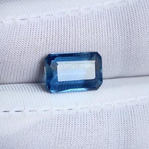 May include: A rectangular, faceted gemstone with a deep blue hue and a lighter, rectangular inclusion in the center. The gem is set against a white, textured background, highlighting its clarity and color. The cut of the gem is precise, reflecting light.