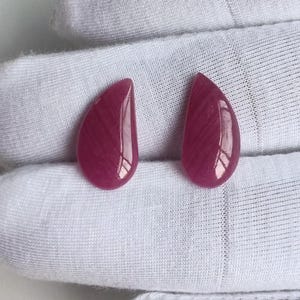May include: Two polished, teardrop-shaped gemstones in a deep magenta hue. The stones have a glossy finish and a subtle, striated pattern. They are displayed on a white, textured surface.