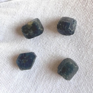 May include: Four small, rough-cut, dark blue sapphire gemstones arranged in a square formation. Each stone features an irregular, multifaceted shape with deep navy and slate blue tones, showcasing natural textures and varied surface light reflections.