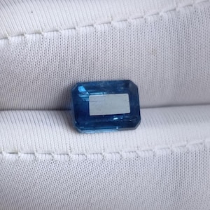 May include: A rectangular, faceted, blue gemstone with a light-colored rectangular inclusion. The gem is set against a white, textured background. The gemstone's facets reflect light, creating a sparkling effect. This gem could be used in jewelry.