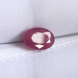 May include: Oval-cut ruby gemstone with a deep red hue. The gemstone has multiple facets that reflect light, creating a sparkling effect. The ruby is set against a white background.