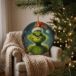 May include: A round ceramic Christmas ornament featuring a detailed illustration of a green, furry creature with a mischievous expression wearing a red and white Santa hat, suspended by a gold string against a dark, festive background.