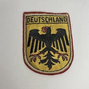 Puede incluir: Un parche bordado en forma de escudo que muestra un águila negra con pico y garras rojas sobre un fondo dorado. La palabra DEUTSCHLAND está bordada en negro en la parte superior. El parche tiene un borde color crema y un remate rojo.
