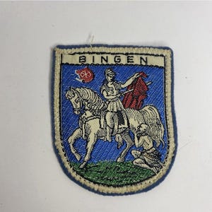 以下が含まれることがあります： 上部にBINGENの文字が入った盾形の刺繍ワッペン。青い背景に、ヘルメットと赤いマントを身につけ白い馬に乗った人物と、緑の地面にひざまずく小さな人物が描かれています。