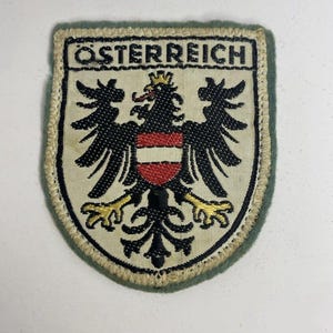 May include: A shield-shaped embroidered patch featuring the text OSTERREICH at the top. The design displays a black eagle with a gold crown and yellow talons, centred with a red and white horizontal striped shield on a cream-coloured background.