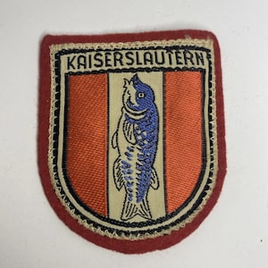 以下が含まれることがあります： 下部の角が丸い長方形の刺繍ワッペンで、赤い縁取りが施されています。中央にはベージュの縦縞があり、その両側はオレンジ色です。ベージュの縞模様の中央には青い魚が配置され、上部にはKAISERSLAUTERNという文字が刺繍されています。