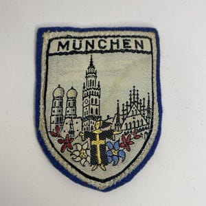 May include: A shield-shaped embroidered patch with a blue border and a cream-colored center. It features a black-stitched cityscape of Munich, including the Frauenkirche, with the text MUNCHEN at the top and colorful floral accents at the base.