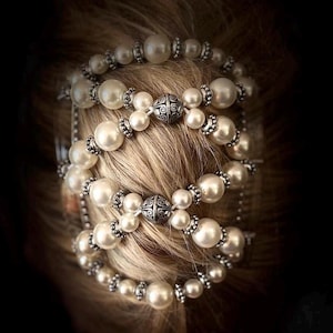 May include: A pearl hair accessory with multiple strands of large, round, white pearls. The pearls are interspersed with silver-colored beads and decorative metal accents. The accessory is set against a backdrop of blonde hair.