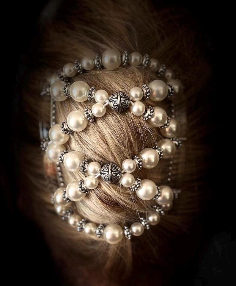 May include: A pearl hair accessory with multiple strands of large, round, white pearls. The pearls are interspersed with silver-colored beads and decorative metal accents. The accessory is set against a backdrop of blonde hair.