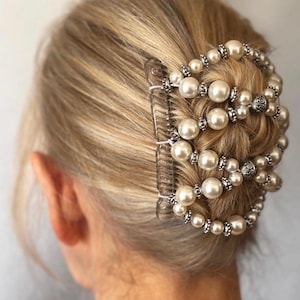 May include: A clear hair comb with a decorative design of faux pearls and silver-colored beads. The comb is set in blonde hair, creating a formal updo. The pearls are large and round, and the beads are small and sparkly.