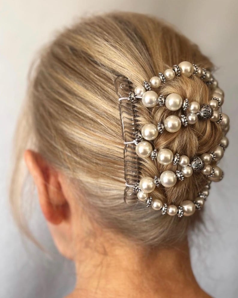 May include: A clear hair comb with a decorative design of faux pearls and silver-colored beads. The comb is set in blonde hair, creating a formal updo. The pearls are large and round, and the beads are small and sparkly.