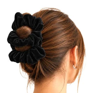 May include: A black velvet hair scrunchie with a gathered, ruffled texture. The fabric has a soft, matte appearance and is wrapped around a bun of brown hair. The accessory features a circular, elasticised design that creates volume and folds.