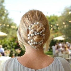 May include: A woman's blonde hair is styled in a low bun, decorated with a pearl and silver beaded hair accessory. The hairpiece is the focal point, adding elegance. The background is blurred, suggesting an outdoor event.