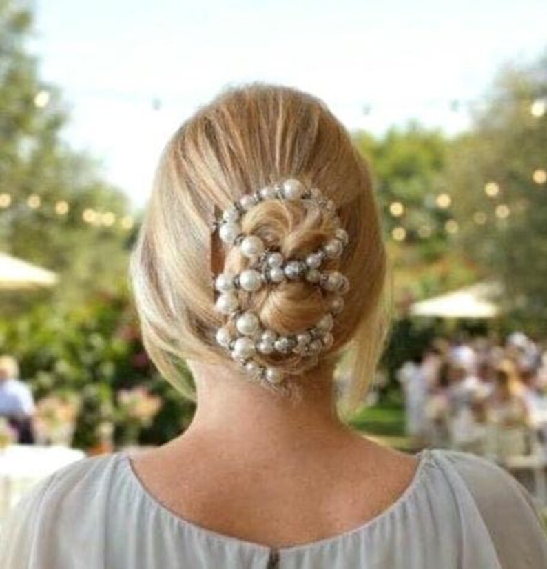 May include: A woman's blonde hair is styled in a low bun, adorned with a pearl and silver beaded hair accessory. The hairpiece is the focal point, adding elegance. The background is blurred, suggesting an outdoor event.