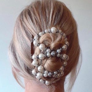May include: A hairstyle featuring a decorative hair accessory. The accessory is composed of multiple strands of white faux pearls and silver-toned metal beads. The hair is blonde and styled in a bun, with the accessory wrapped around it.