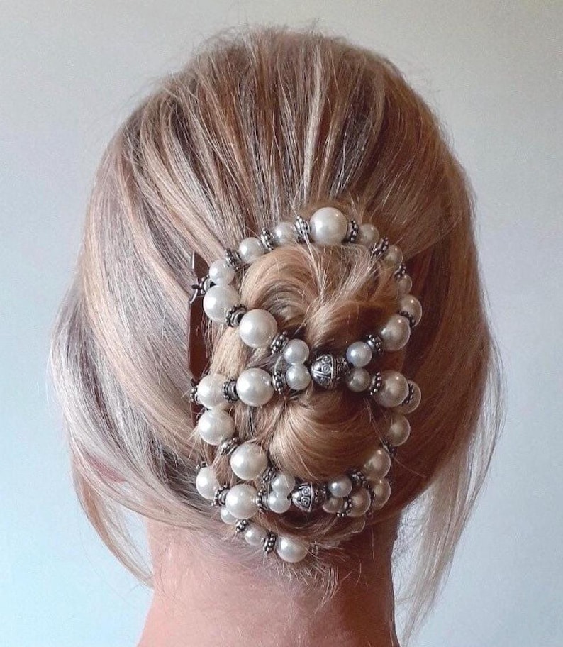May include: A hairstyle featuring a decorative hair accessory. The accessory is composed of multiple strands of white faux pearls and silver-toned metal beads. The hair is blonde and styled in a bun, with the accessory wrapped around it.
