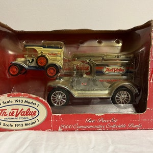 Puede incluir: Dos bancos de colección de modelo T de 1913 de estilo vintage, uno en dorado con la marca "True Value Hardware Stores" y el otro en blanco con detalles azules. Los bancos se muestran en una caja roja y blanca con el texto "1:43 Scale 1913 Model T" y "1:25 Scale 1913 Model T".