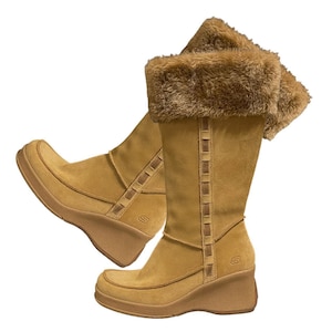 Pode incluir: Botas de camurça castanhas com salto cunha e acabamento em pele sintética. As botas têm um design até ao joelho com costuras decorativas e o logótipo da marca. Sola plataforma e bico arredondado.
