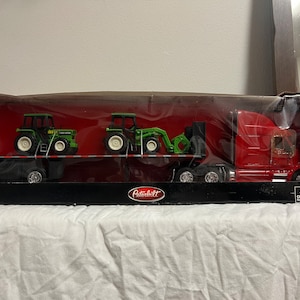 Puede incluir: Un camión semi-remolque Peterbilt rojo de fundición a presión con una parte trasera abatible que transporta dos tractores John Deere verdes en un remolque de plataforma. La caja dice "1:32 Die-Cast Openable Back" y "NewRay".
